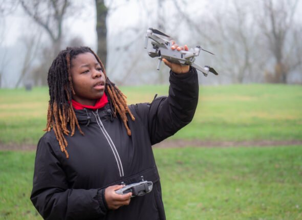 Young african girl standing checking flying drone camera outdoors in a park in cold weather.